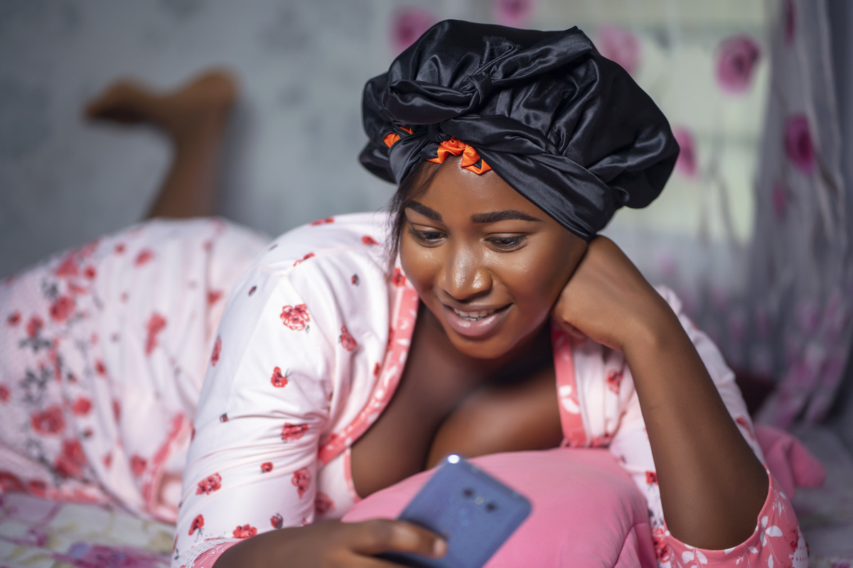 Portrait of a Young African American Female in a Satin Bonnet Using Her Phone on Her Bed